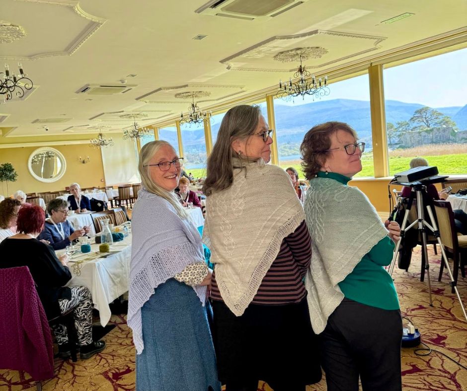 Three women look back over their shoulders to showcase their intricately hand-knitted lace and textured shawls inside a dining room.