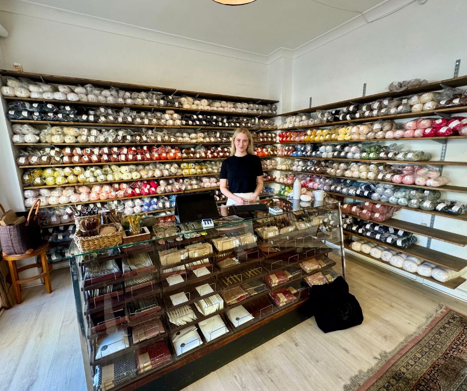 Woman standing behind a glass display counter smiling in a cozy yarn shop filled with wall-to-wall shelves stacked with colorful skeins of yarn and knitting supplies.