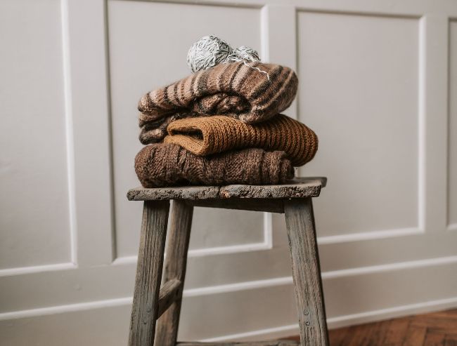 A stack of folded brown and tan knit sweaters topped with a ball of yarn, sitting on a rustic wooden stool.