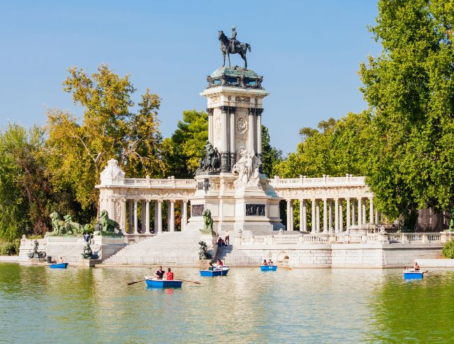 A wide shot of the Monument to Alfonso XII in El Retiro Park, Madrid, featuring a large columned semi-circle structure topped with an equestrian statue, fronted by a large lake where people are riding in small blue rowboats under a clear blue sky.
