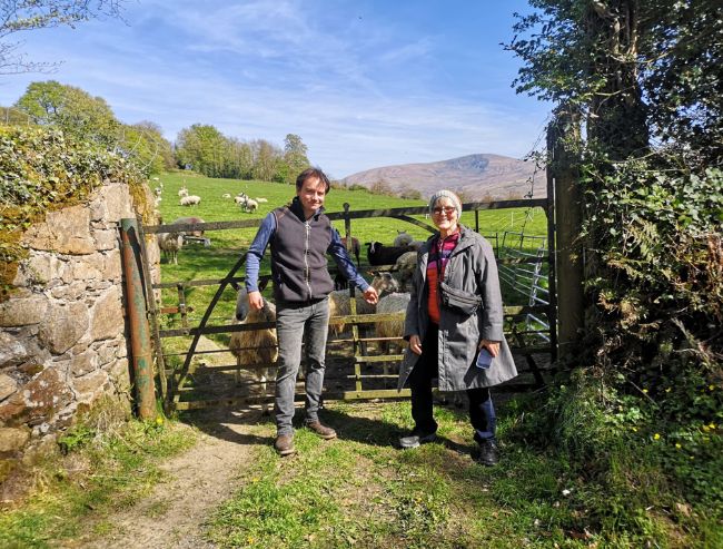 A man and woman stand by an iron farm gate on a sunny day, with sheep grazing in a green hilly pasture and mountains behind them.