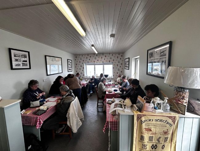 A group of people sit at long tables with red-and-white checkered cloths inside a narrow room, knitting and working with yarn together.