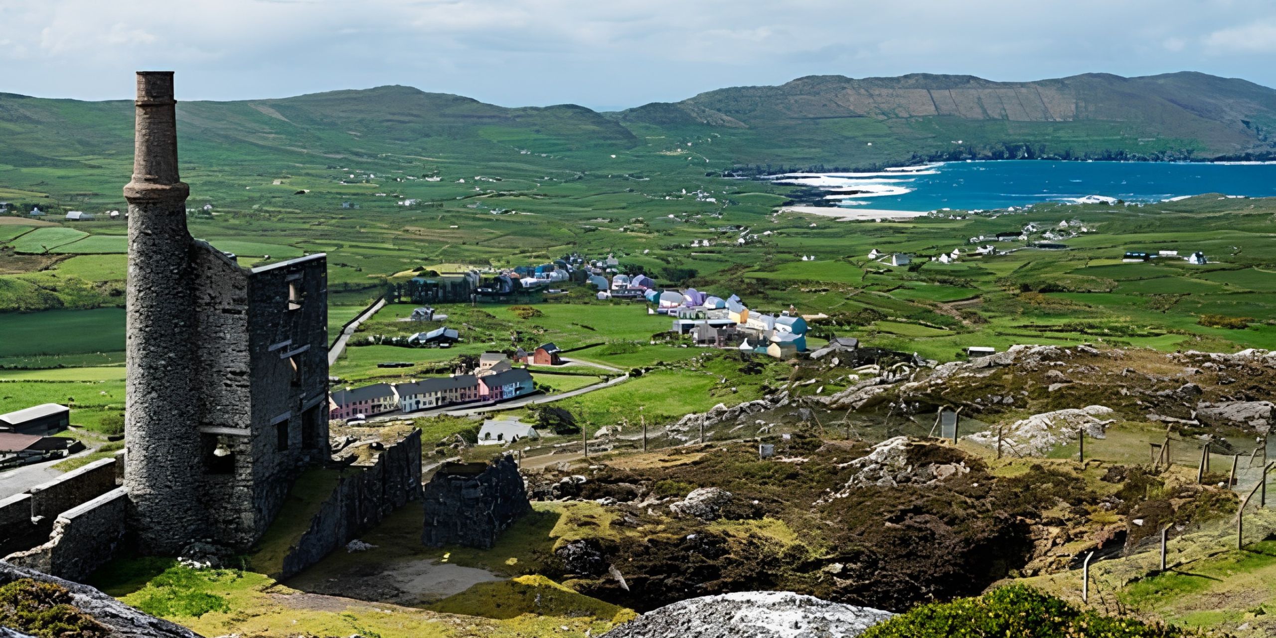 Panoramic view of the Dingle Peninsula with the historic Ballycorick Copper Mine chimney overlooking green fields, a coastal bay and rolling hills in County Kerry, Ireland.