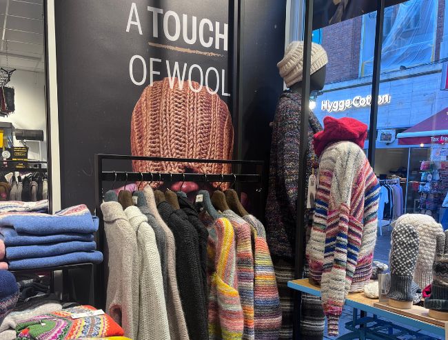 A store display featuring rows of colorful knit sweaters on a clothing rack beneath a large sign. 