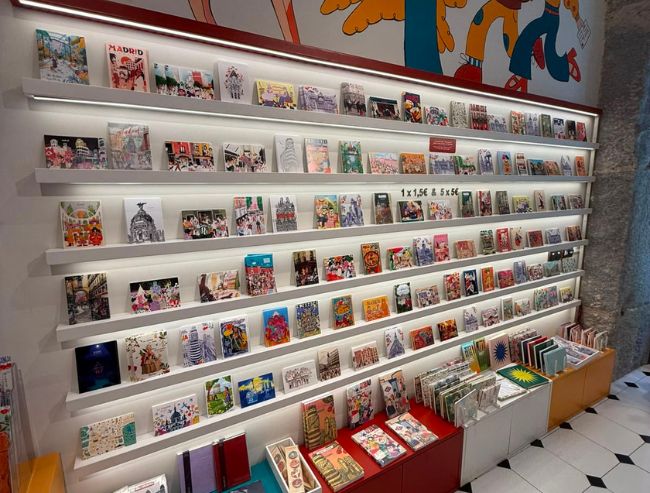 A wall inside La Postalera souvenir shop lined with multiple rows of thin white ledges displaying a large, colorful variety of artistic postcards, mini prints and illustrated notebooks.