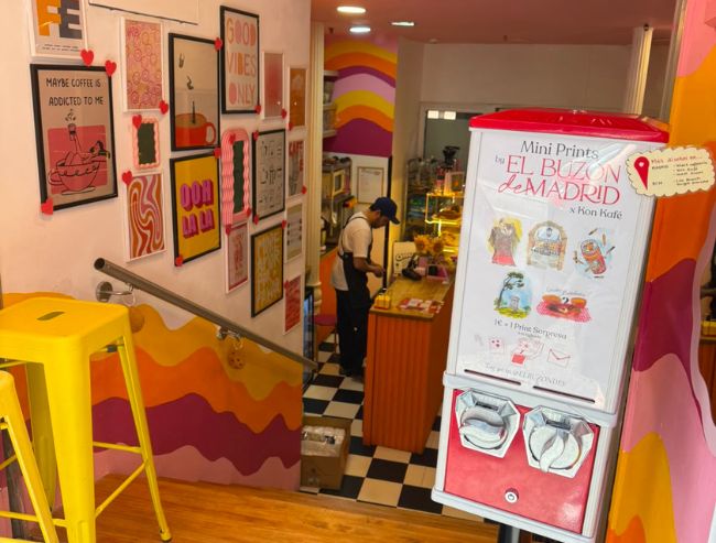 A view looking down a wooden staircase with a bright yellow metal stool on the left, leading into a colorful café with wavy pink, orange, and yellow painted walls, a checkered floor, and a red "El Buzón de Madrid" mini-print vending machine in the foreground.