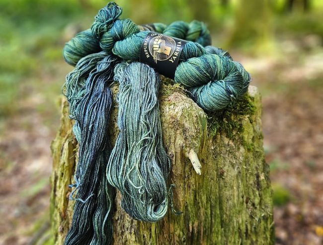 A skein of blue-green hand-dyed yarn draped over a moss-covered tree stump outdoors.