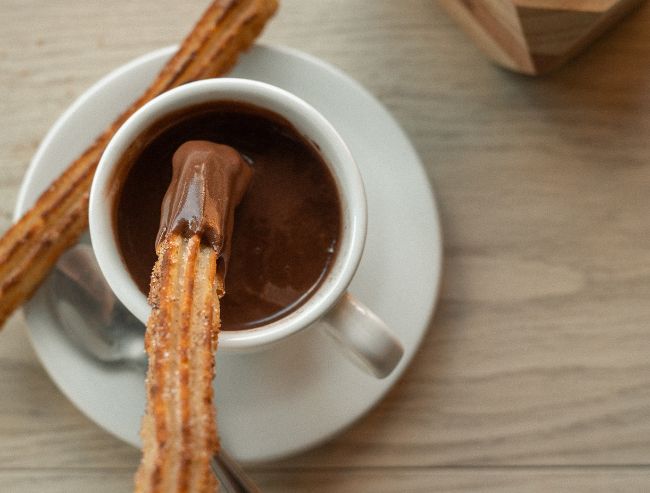 A close-up, top-down view of a long, golden-brown churro being dipped into a small white ceramic cup filled with thick, dark hot chocolate sitting on a white saucer.