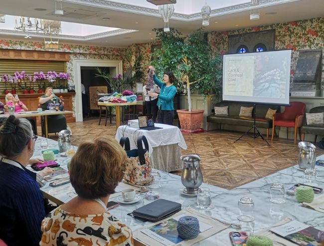 People attending a yarn and knitting workshop in an open room, with yarn, craft materials and a presentation screen on display.