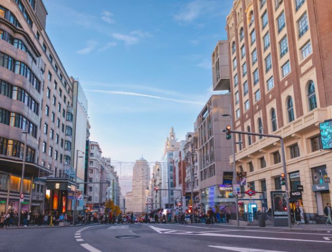A ground-level street view looking down the wide, curved avenue of Gran Vía in Madrid, lined with grand multi-story architecture, city traffic lights, and pedestrians on the sidewalks under a blue sky with light clouds.