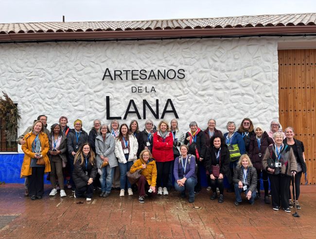 A large group of people poses for a photo together in front of a textured white wall with large black text reading "ARTESANOS DE LA LANA."