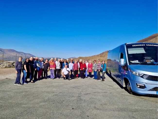 A group of tourists posing beside a blue coach bus in a mountainous landscape under a clear blue sky.