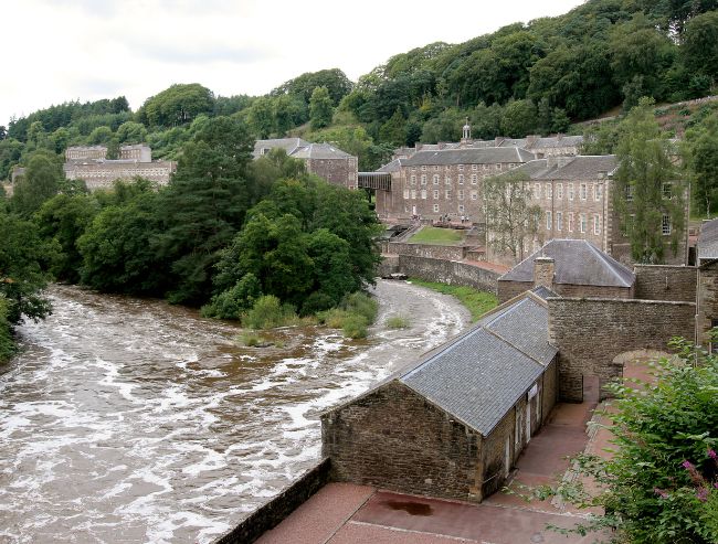 A historic stone mill complex beside a fast-flowing river, surrounded by lush green woodland.