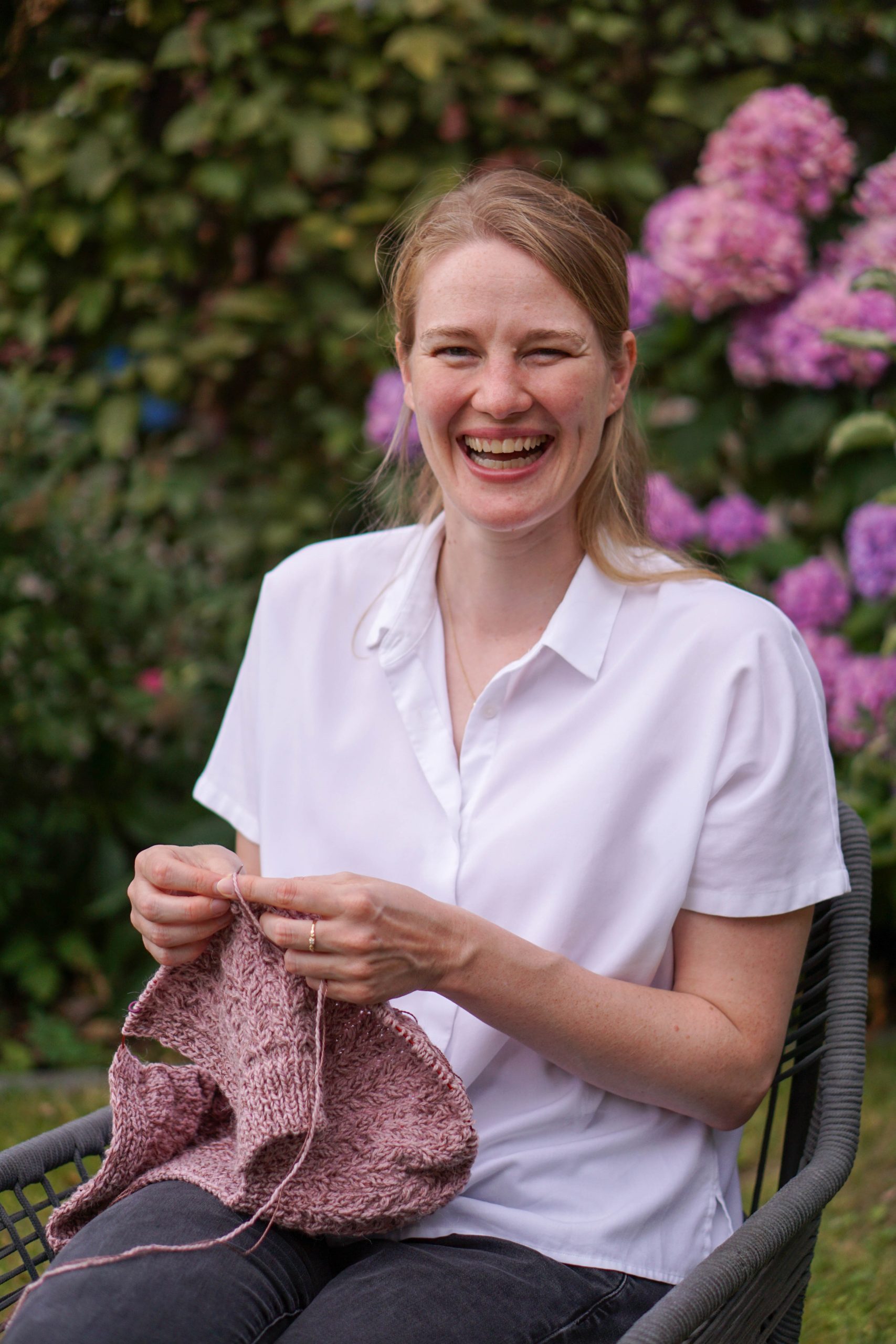 Woman knitting pink yarn with pink flowers and green bush in the background.