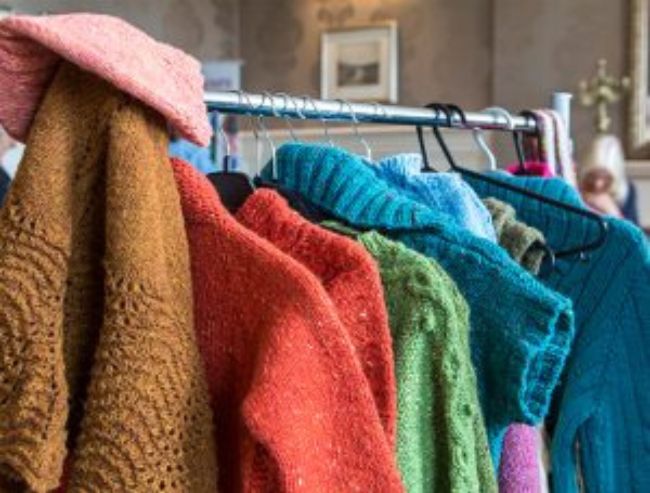 A clothing rack holds a row of colorful, hand-knitted woolen garments, including sweaters and cardigans in brown, orange, green, and blue.