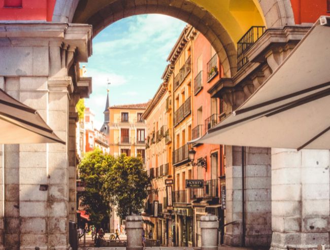 A massive stone archway of Plaza Mayor in Madrid, framing a picturesque street outside with warm orange and yellow European-style buildings under a bright sky.