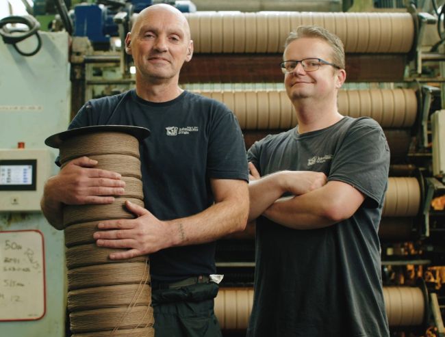 Two male textile workers posing inside a mill, with one worker holding a large spool of brown yarn and the other standing with arms crossed.