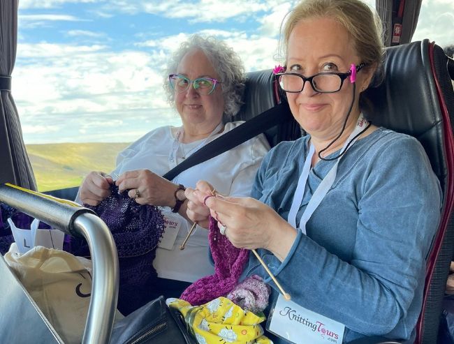 Two smiling women sit next to each other on a tour bus, both holding knitting needles and actively working on colorful yarn projects.