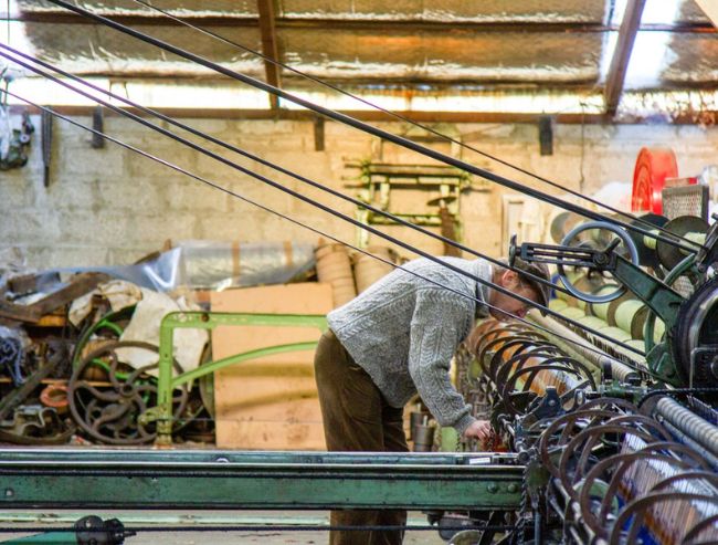A man leaning over a complex, vintage mechanical weaving loom inside a rustic workshop or mill.