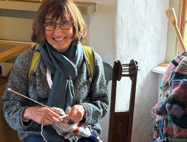 A smiling woman with short brown hair, wearing a gray sweater and blue scarf, knits with light-colored yarn while seated indoors near textile equipment.