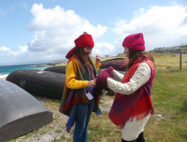 Two women in colorful knitwear, hats, and ponchos standing by a grassy shoreline with overturned boats.