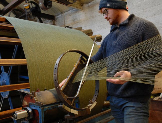 A man wearing a beanie and glasses operates a large textile machine, guiding a dense sheet of parallel green threads.