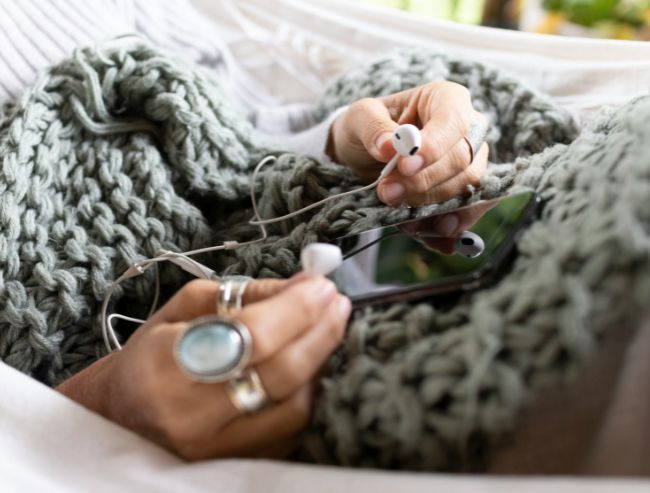 A close-up of a person under a chunky green knit blanket holding wired earphones and a smartphone.