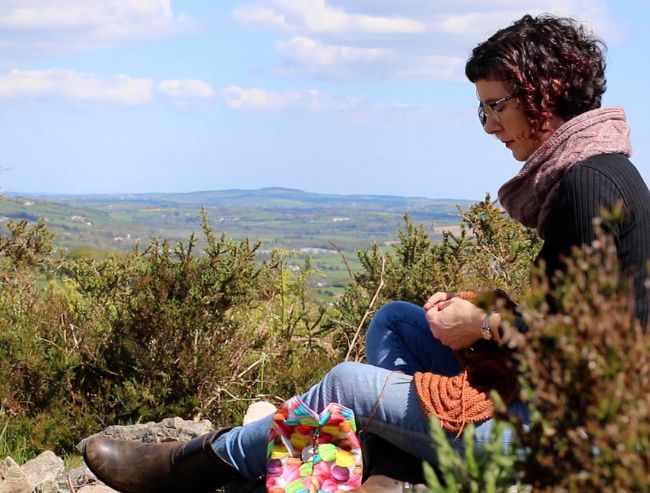 A woman sitting on a rocky hillside knitting with orange yarn, overlooking a vast green valley.