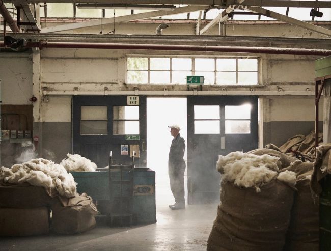 A worker standing by an open doorway in a large industrial warehouse, surrounded by large burlap sacks filled with raw white wool.