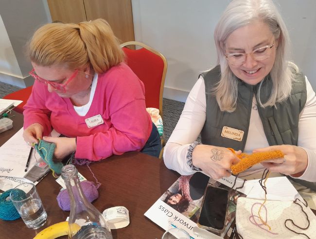 Two women sitting at a table during a knitting workshop, working with teal and yellow yarn.