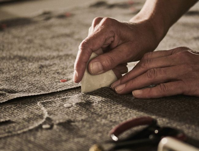 A close-up of a pair of hands using a piece of tailor's chalk to mark a piece of textured grey fabric on a worktable.