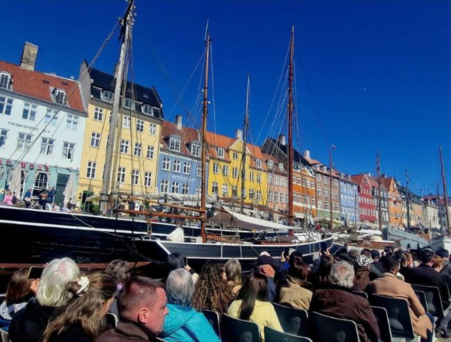 A group of people on a boat tour look toward a waterfront lined with historic, brightly colored buildings and tall masted ships under a clear blue sky.