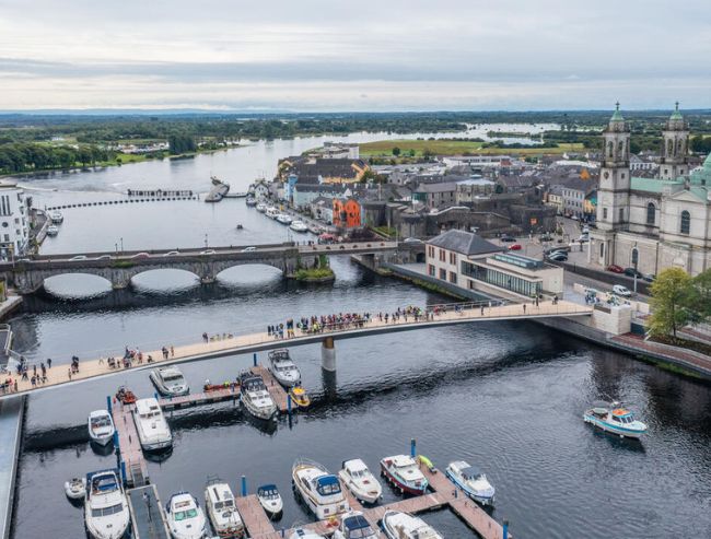 An aerial view of a river flowing through a town, featuring two parallel bridges, a small boat marina and a large stone church with dual steeples.