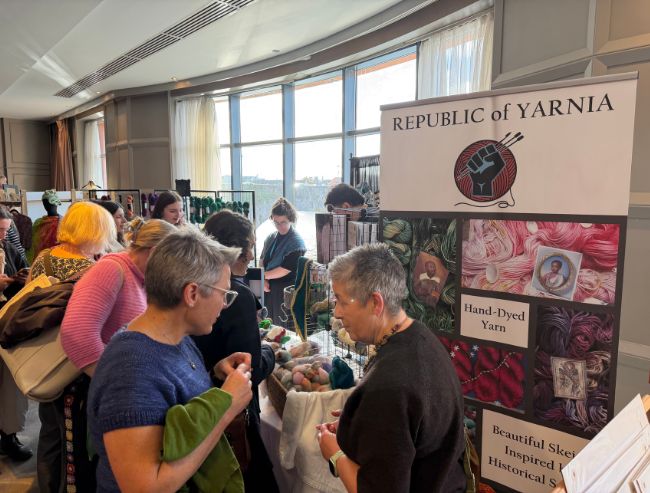 A crowded indoor market stall for "Republic of Yarnia," where attendees browse baskets of hand-dyed yarn.