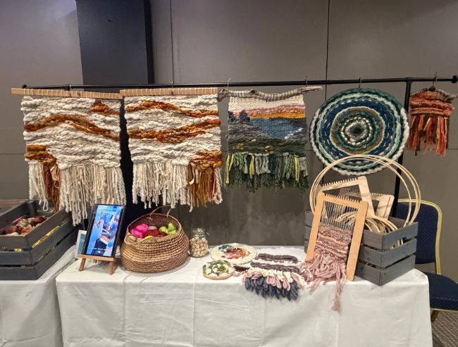 A craft display table featuring woven wall hangings in earth tones, smaller circular textiles and small hand looms in crates.