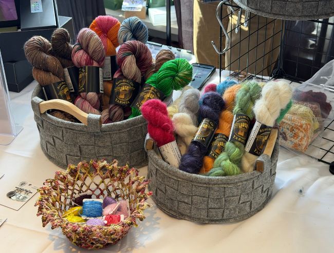 Two grey felt baskets filled with vibrant, multi-colored skeins of hand-dyed yarn on a white tabletop display.